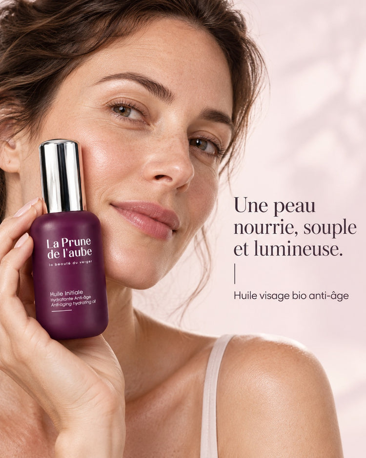 Femme tenant le flacon de l’Huile Initiale près de son visage. Une peau nourrie, souple et lumineuse. — Huile visage bio anti-âge