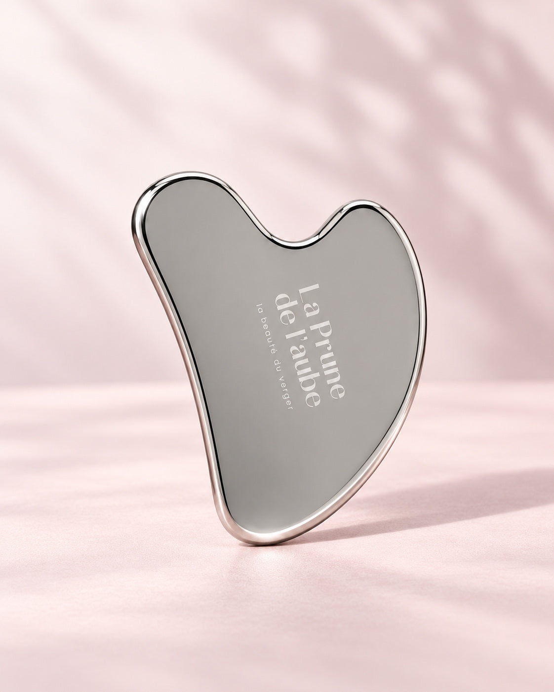 Gua Sha en acier inoxydable La Prune de l’aube sur un fond rose.
