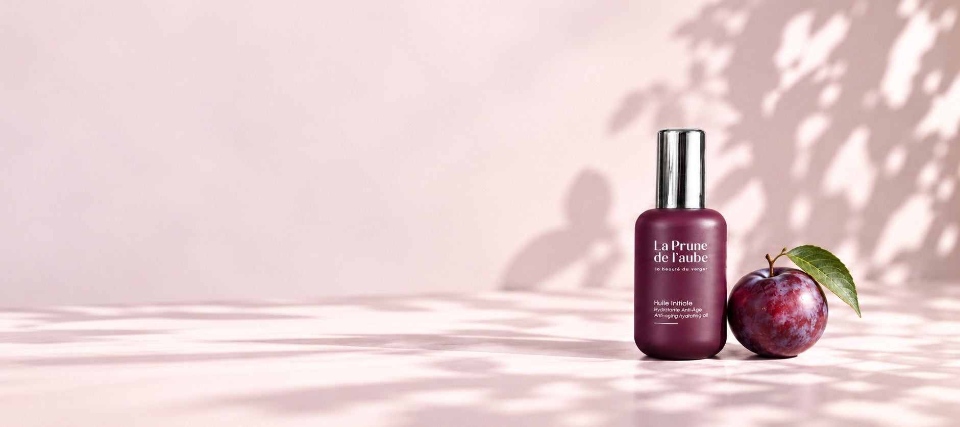 Flacon de L’Huile Initiale accompagné d’une prune sur un fond rose lumineux.